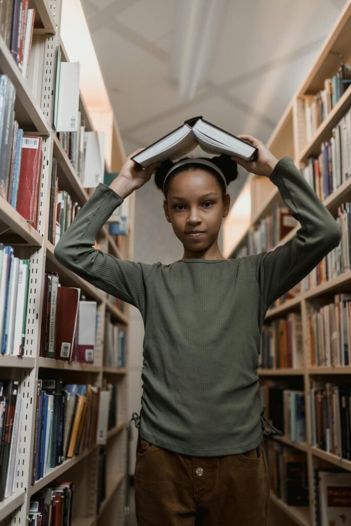 A young girl in a library balancing books on her head with bookshelves on either side.